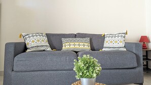 A grey sofa with patterned cushions, a potted plant, and a wall art piece above. - Apartment close to port, beaches, golf course and ile des Loisirs with air conditioning (Agde)