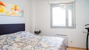 A bedroom with a bed, a window with a white valance, a radiator, and a wall-mounted chair.