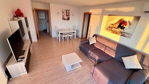 A modern living room with a flat-screen TV, a sofa, a coffee table, and a dining area with chairs.