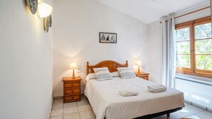 A bedroom with a wooden bed, two bedside tables with lamps, a small dresser, a window with curtains, and a wall-mounted picture. - Villa Flor, relax in Menorca (Cap d`Artrutx)