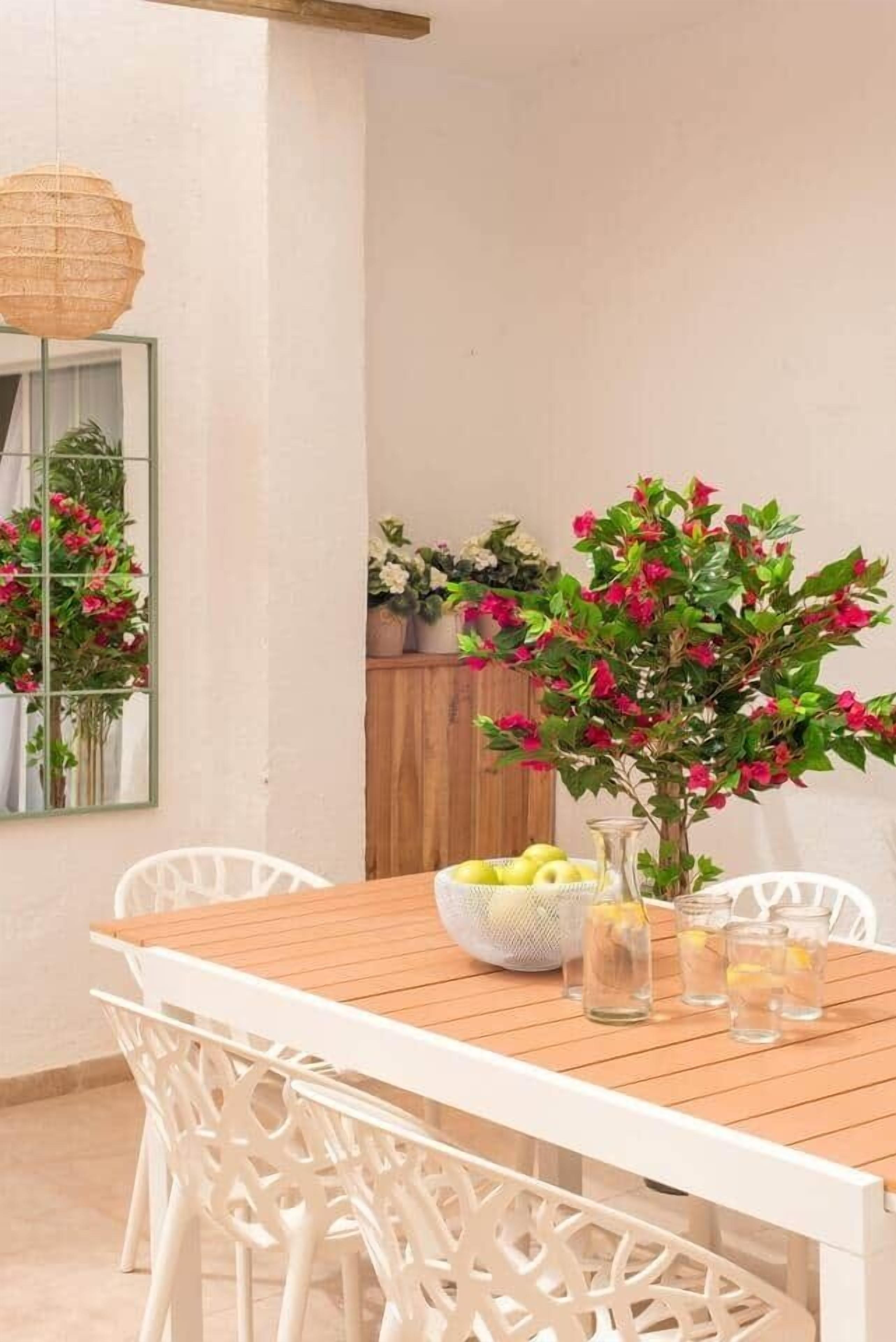 Una mesa de madera con sillas blancas, un jarrón con flores rojas y un cuenco de limones.