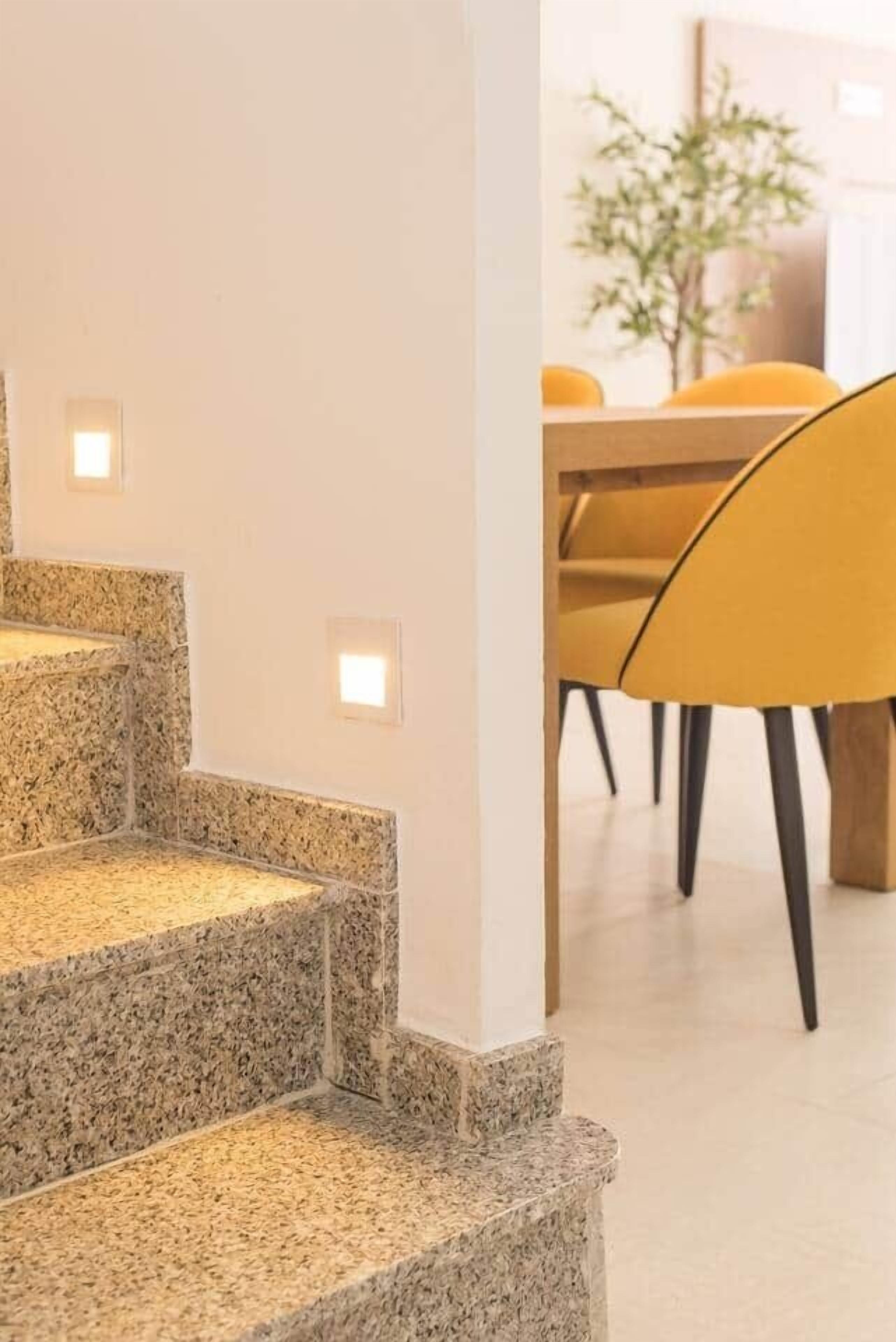Een trap met granieten treden en inbouwverlichting, die leidt naar een ruimte met een houten tafel en gele stoelen.