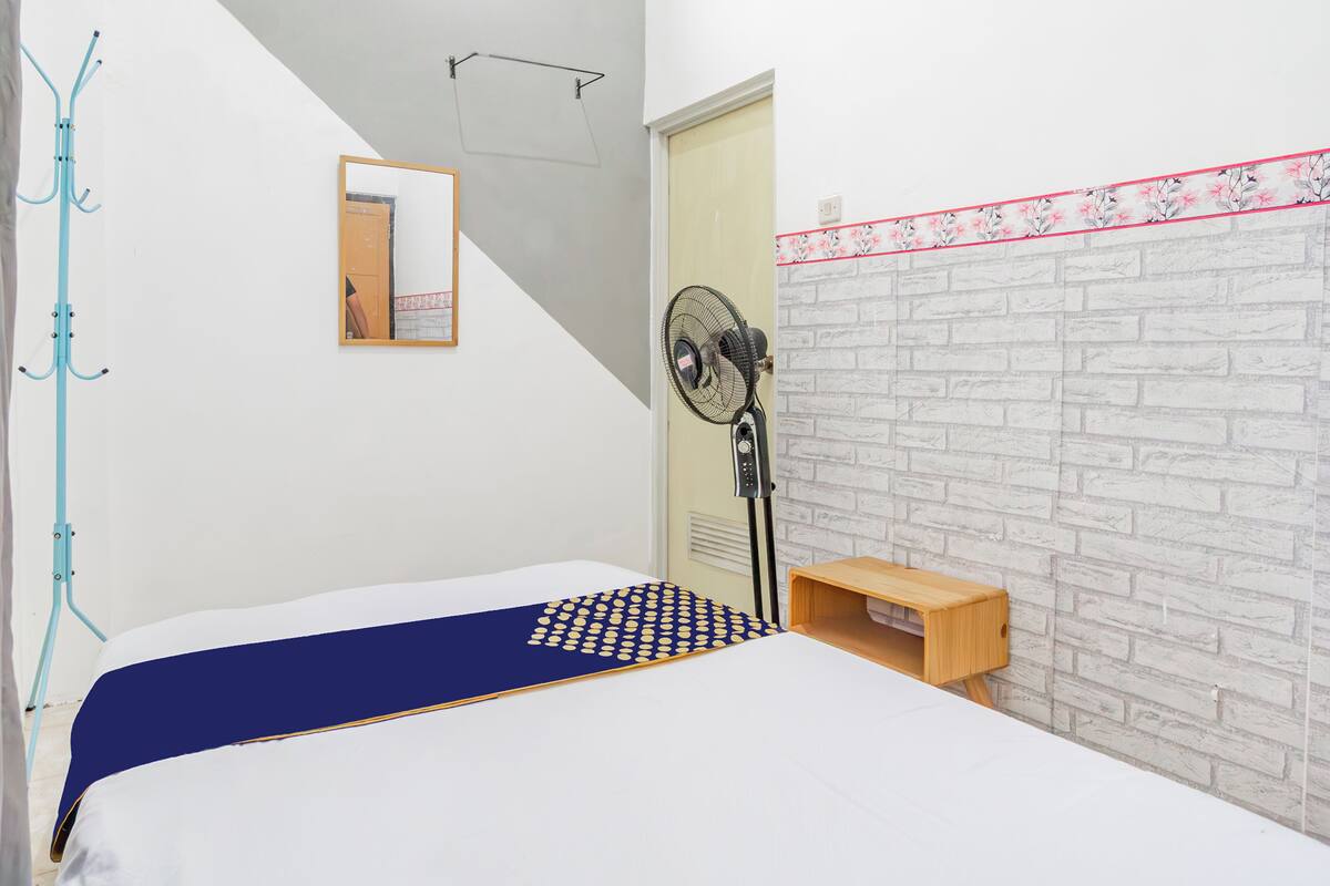 A bedroom with a bed, a fan, a coat rack, and a small wooden shelf.