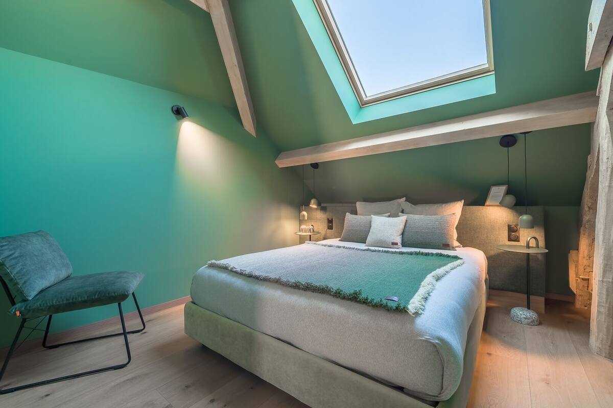 Une chambre avec un puits de lumière, un lit, une chaise et une table de chevet avec une lampe.