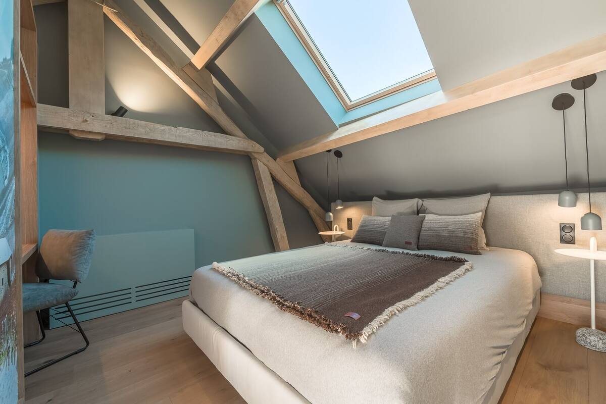 Une chambre aménagée dans les combles, avec un puits de lumière, un lit recouvert d’un plaid, une chaise et des lampes suspendues.