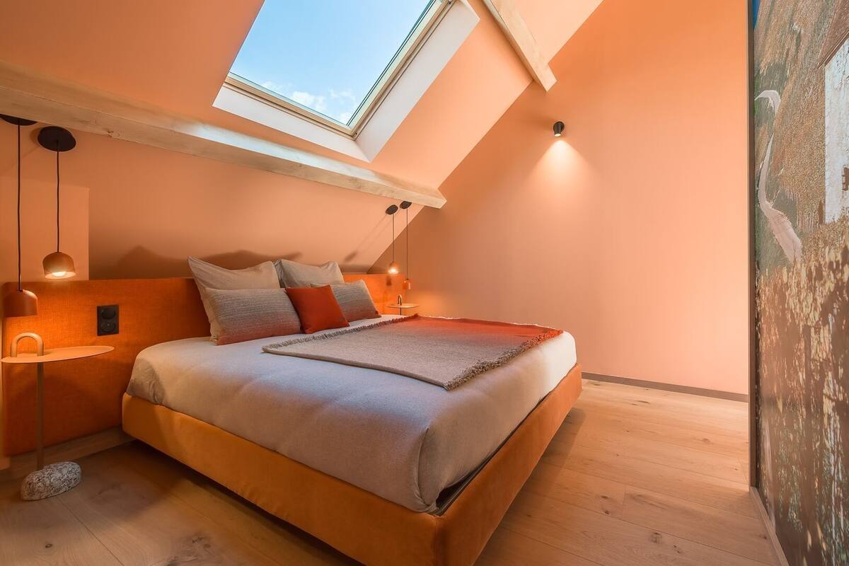 Une chambre avec un puits de lumière, un lit avec des oreillers, une table de chevet et une lampe fixée au mur.