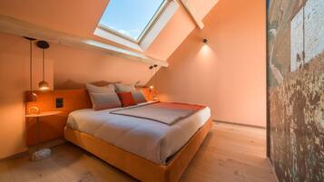 Un dormitorio con tragaluces, una cama con almohadas, una mesita de noche y una lámpara fijada a la pared.