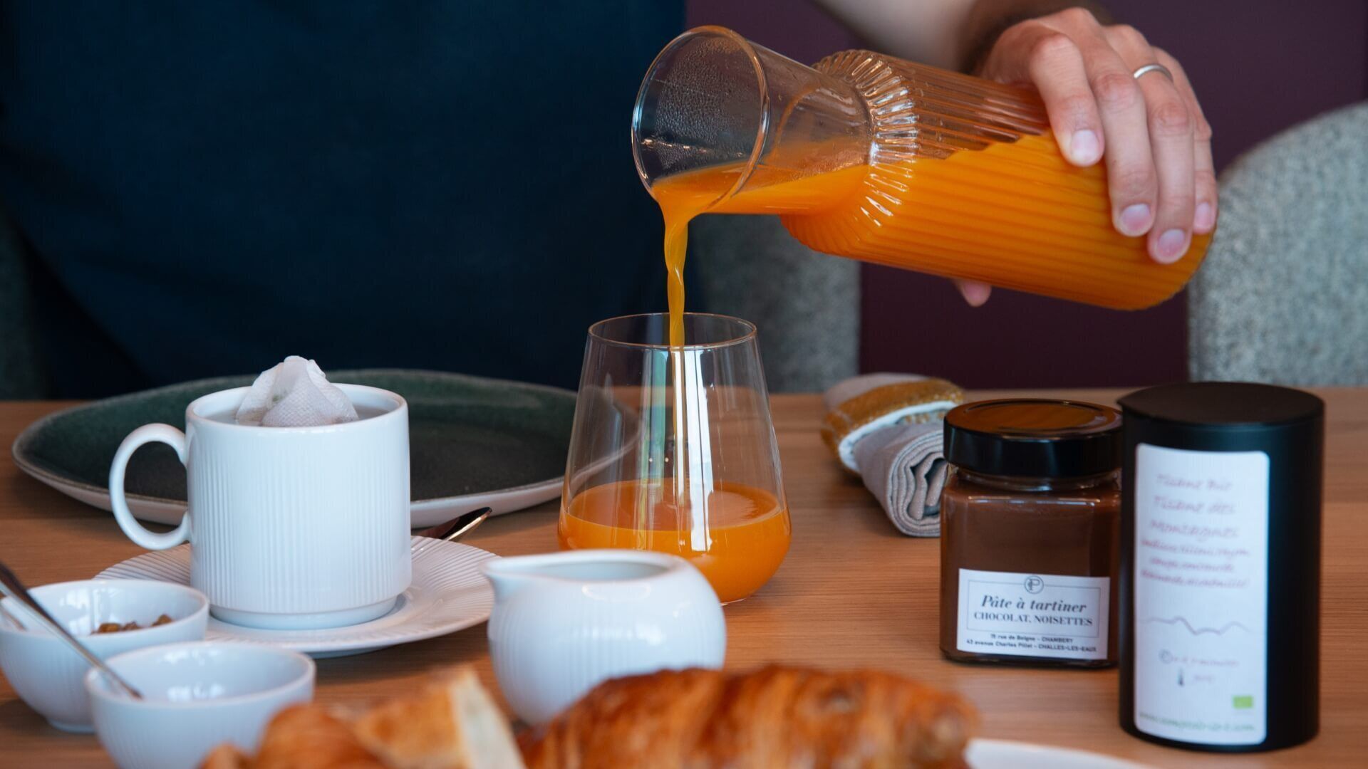Ein Frühstückstisch mit Orangensaftgläser, einer Kaffeetasse, einem Schokoladenaufstrich und einem Croissant.