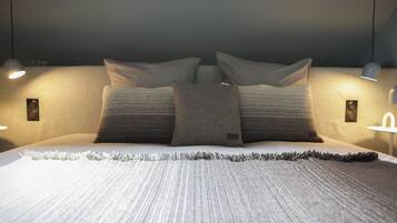 Een net opgemaakt bed met een deken met structuur, aan weerszijden met wandlampen en wandspots.