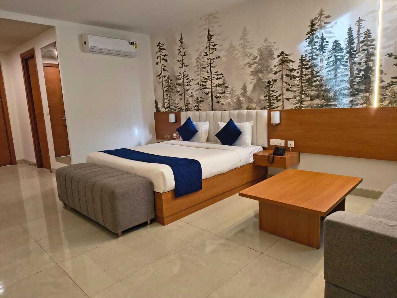 Una habitación de hotel moderna con una cama grande, una mesita de noche, una mesita de madera, un sillón gris y un ventanal con un mural de un bosque.