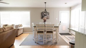 Un salon avec un canapé beige, une table à manger avec quatre chaises, un lustre et une grande fenêtre avec des stores.