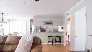 Un salon moderne avec un canapé en cuir marron, un coussin blanc et une table basse en bois. Au fond, on aperçoit un coin cuisine avec un comptoir blanc et deux tabourets de bar.