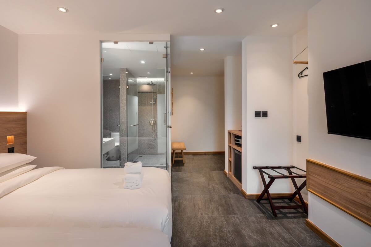 Una habitación de hotel moderna con una cama grande, un televisor montado en la pared, zona de ducha y un pequeño rincón de descanso.