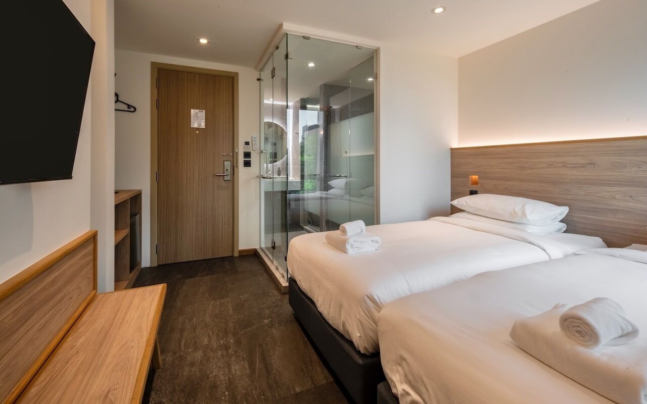 Une chambre d’hôtel avec deux lits, un banc en bois, une télévision et une douche séparée par une paroi en verre.