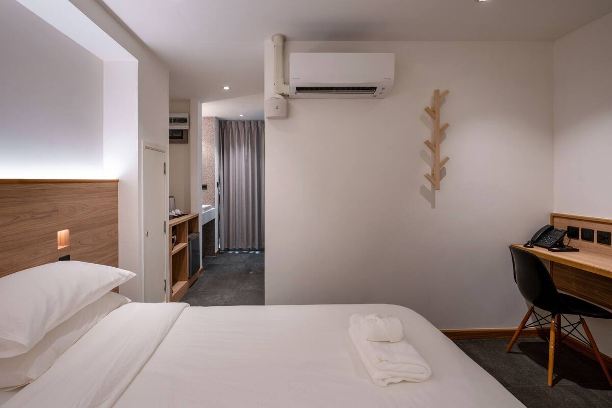 Habitación de hotel con cama, escritorio, silla, unidad de aire acondicionado y baño visible a través de una puerta abierta.