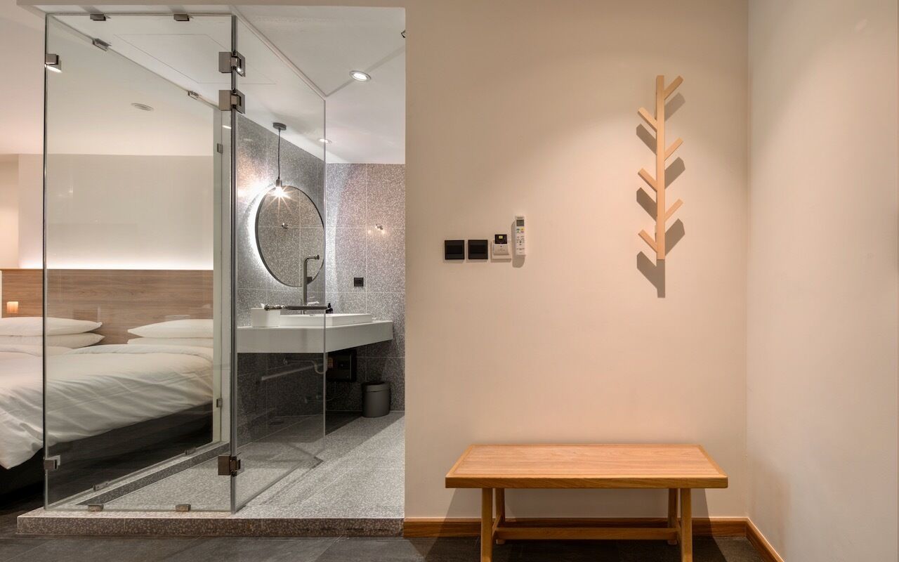 A modern hotel room with a glass-enclosed shower, a wooden bench, and a minimalist decor including a wall-mounted light fixture and a wooden leaf design.