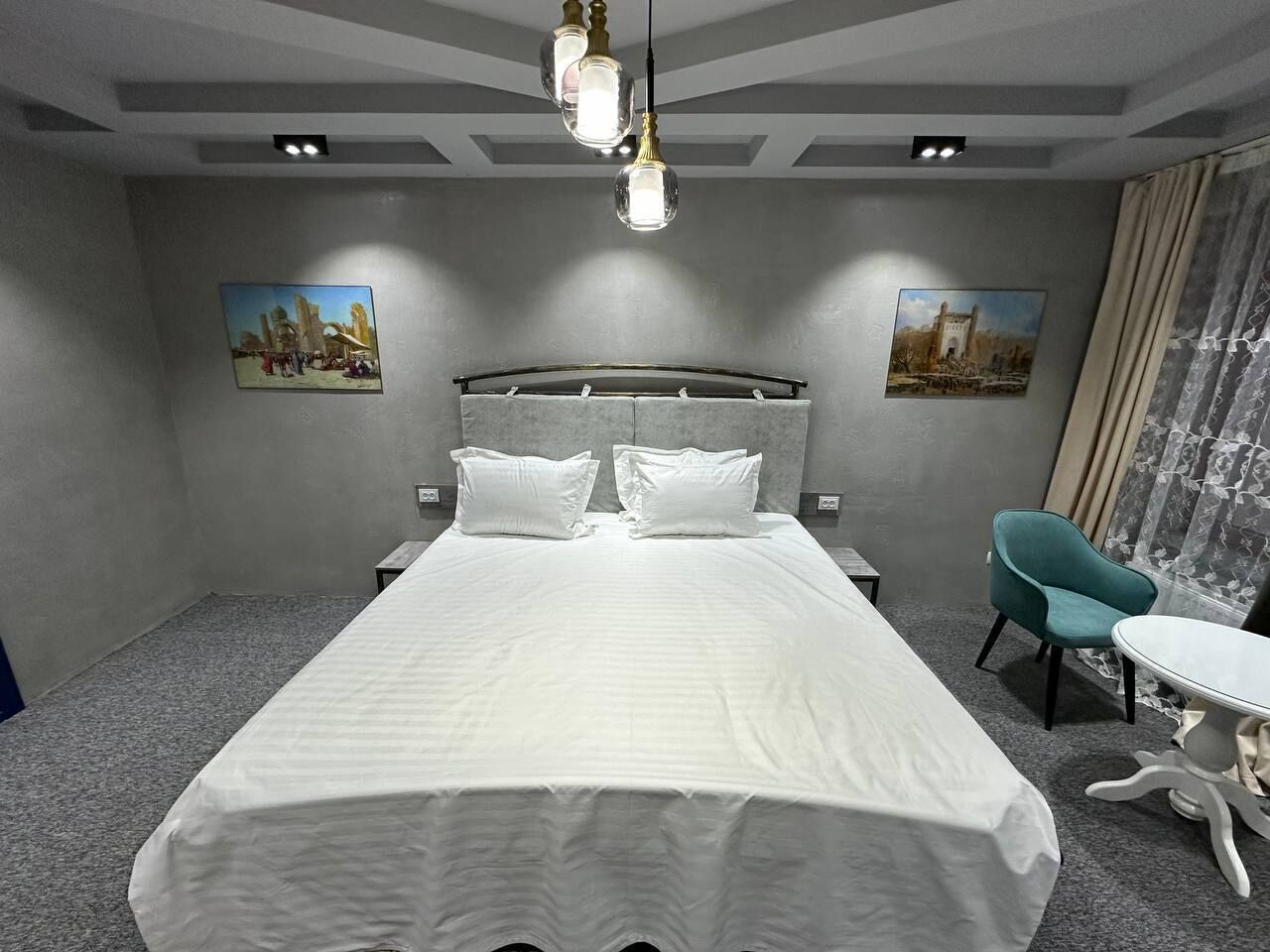 A modern hotel room with a large bed, a small table, a chair, and two wall-mounted paintings.