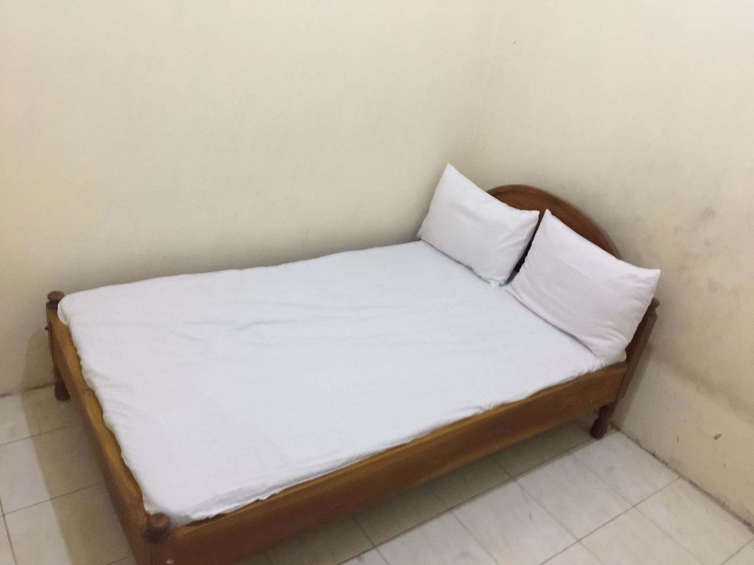 Una cama individual con estructura de madera y ropa de cama blanca en una habitación con paredes lisas y suelo de baldosas.