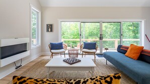A modern living room with a TV, a sofa, two chairs, a coffee table, and a large sliding glass door leading to an outdoor deck. - Airy Tree Canopy Home w/ Creek Views & Family Suite (Sparrow Bush)