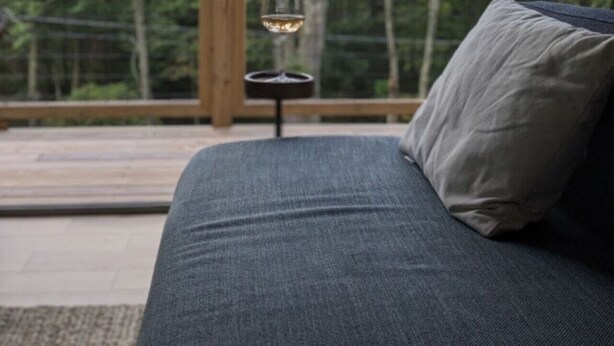 A sofa with a cushion, a small table with a candle, and a view of trees through a wooden railing. - Airy Tree Canopy Home w/ Creek Views & Family Suite (Sparrow Bush)
