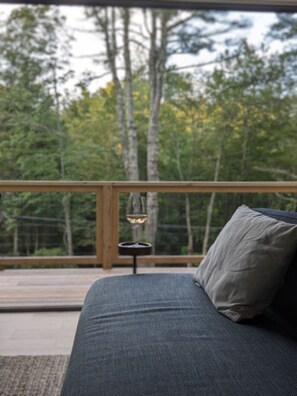 A sofa with a cushion, a small table with a candle, and a view of trees through a wooden railing. - Airy Tree Canopy Home w/ Creek Views & Family Suite (Sparrow Bush)
