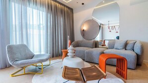 Ein modernes Wohnzimmer mit einem grauen Sofa, einem grauen Sessel und einem weißen Hocker. An der Wand hängt ein großes Spiegelbild und ein runder Tisch mit einer weißen Vase.