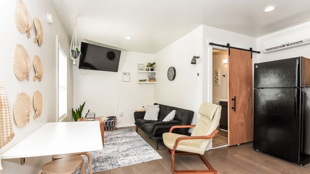 Een knus woongedeelte met een witte tafel, een zwarte bank, een beige fauteuil, een zwarte koelkast en een houten deur.