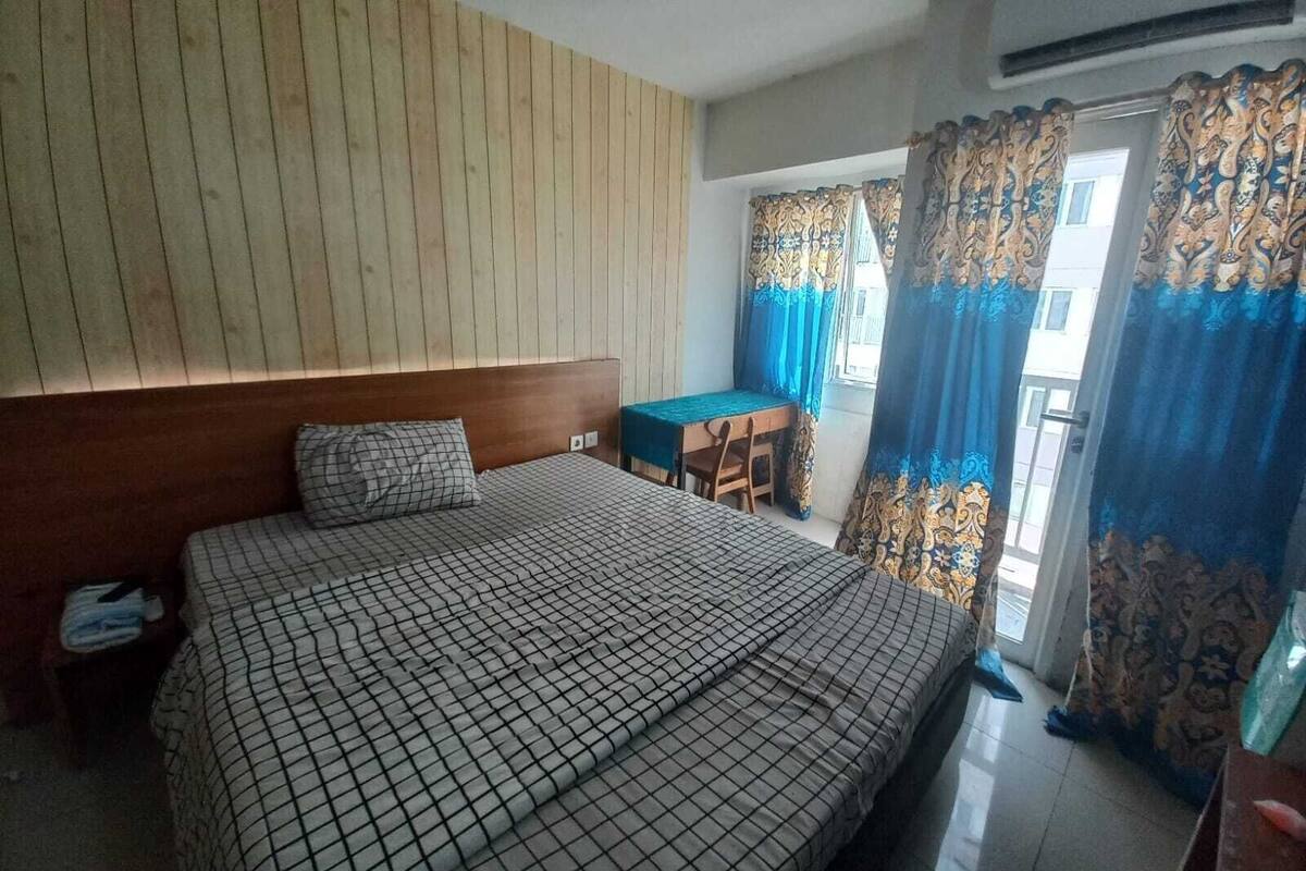 Ein Schlafzimmer mit einem Bett, einem Holz-Kopfteil, einem kleinen Tisch mit Stuhl und einem Balkon mit gemusterten Vorhängen.