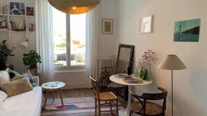 A cozy living room with a sofa, a small round table, and a dining table with chairs. There is a large window with curtains, a bookshelf, and a floor lamp. - Montreuil City, Artists (Montreuil)