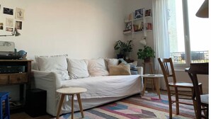 A cozy living room with a white sofa, a small round table, and a colorful rug. There are bookshelves with books and decorative items, and a large window providing natural light. - Montreuil City, Artists (Montreuil)