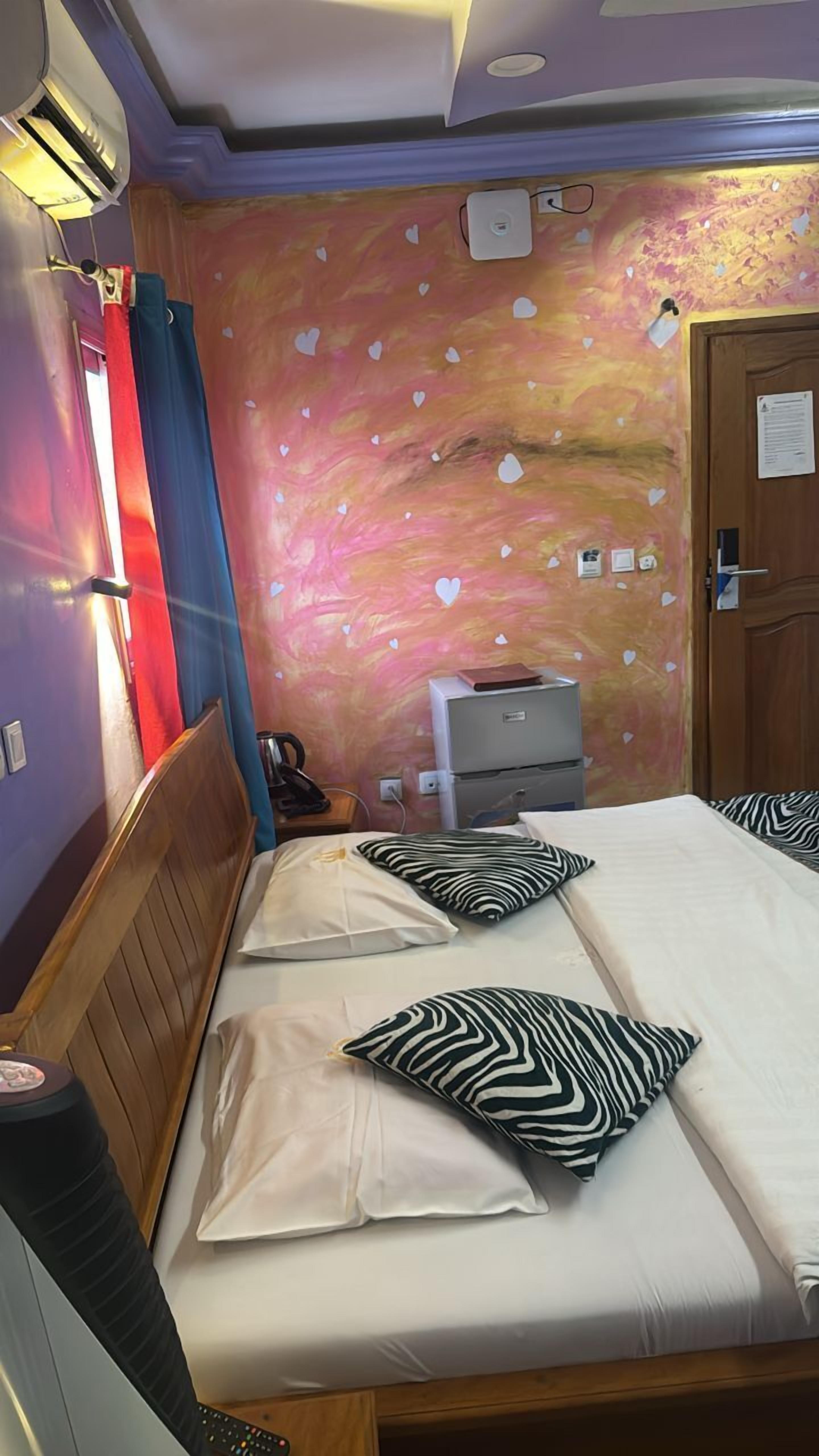 A hotel room with a bed, a nightstand, a dresser, and a wall with a pink and orange abstract design.