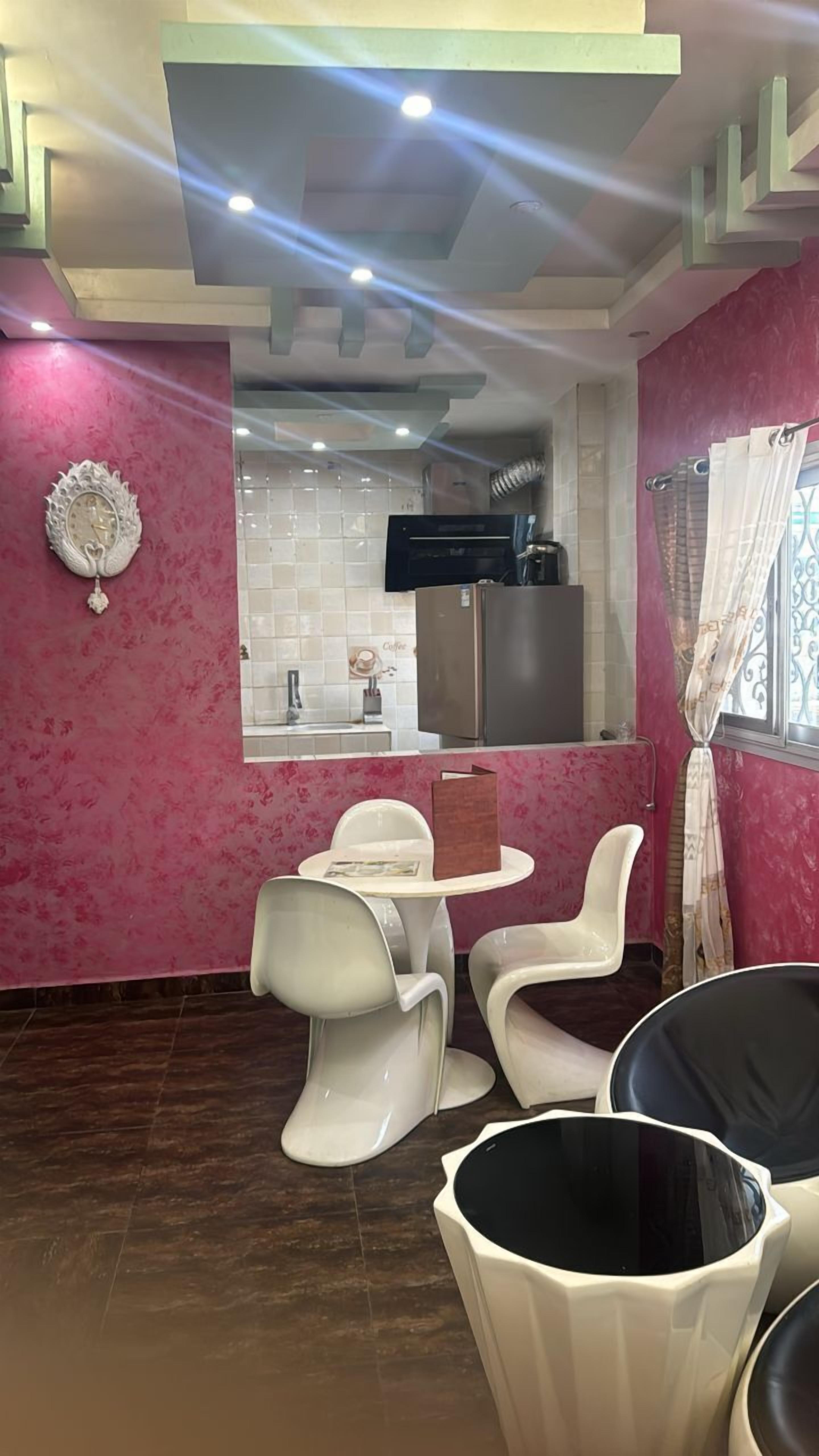 Una cocina moderna con azulejos blancos, un refrigerador y una cafetera. Hay sillas blancas y una mesa en la zona de comedor.