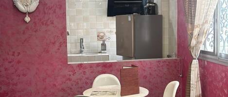 Una cocina moderna con azulejos blancos, un refrigerador y una cafetera. Hay sillas blancas y una mesa en la zona de comedor.
