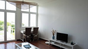 Un soggiorno moderno con una grande finestra, un'area pranzo con sedie in vimini, una TV a schermo piatto su un mobile bianco e un tavolino da caffè in vetro.