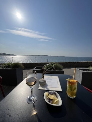 Ein Tisch mit Speisekarte, einem Glas Weißwein, einem Cocktail und einer kleinen Schale Chips und Obst in einem Restaurant am Meer.