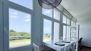 Zona pranzo con tavolo apparecchiato per quattro, ampie finestre che offrono vista su vegetazione e una strada, e un lampadario.