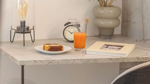 Una mesa de comedor con un plato de comida, un vaso de jugo de naranja, un libro y un florero con plantas secas.