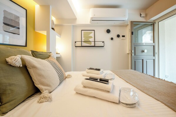 A neatly made bed with towels and a hairdryer, a framed artwork on the wall, and a door with the number 6. - Apt 6 ground floor studio with patio (Athens)