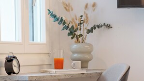 A modern kitchen with a marble countertop, a grey upholstered chair, a vase with dried flowers, a glass of orange juice, and a black alarm clock.