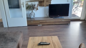 Een woonkamer met een houten salontafel, een bank, een televisie op een houten dressoir en een deur naar een terras.