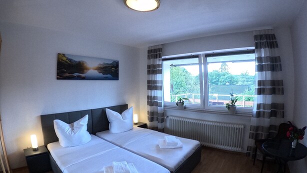 Hotelkamer met twee bedden, een groot raam met gordijnen, een radiator, een kleine tafel met een vaas en een schilderij aan de muur.