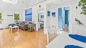 A spacious living room with a large window, a sofa, a coffee table, and a dining area. - Bright & Cozy with Patio, Grill and near the Beach & Golf (Brewster)