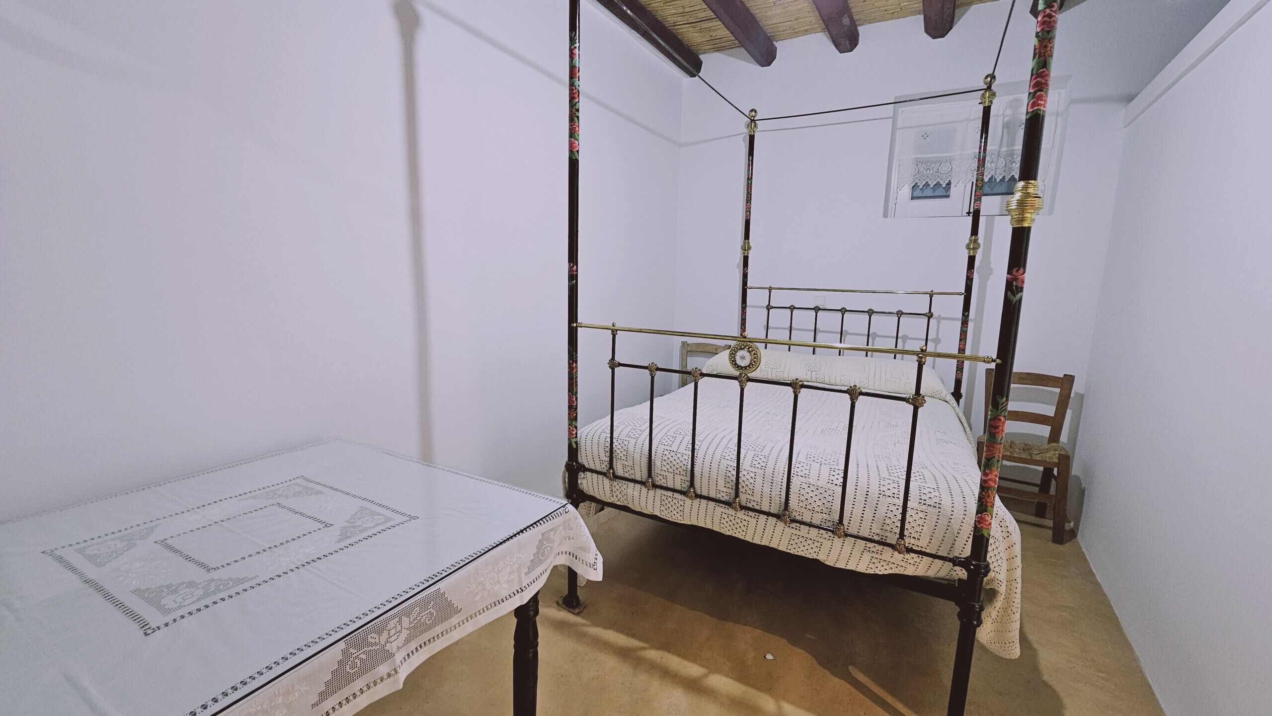 A small, single-bed room with a metal frame, a wooden chair, and a table with a lace tablecloth.