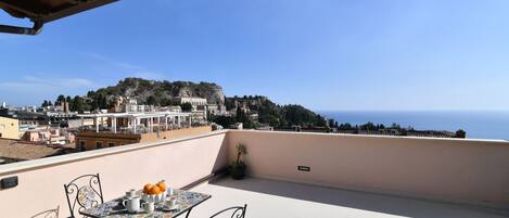 A terrace with a table and chairs, overlooking a coastal town and the sea.