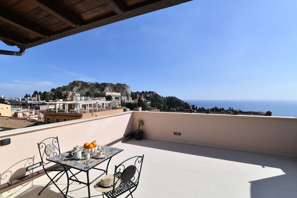 A terrace with a table and chairs, overlooking a coastal town and the sea.