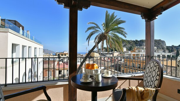 Un balcón con una mesa para dos, con vistas a un paisaje urbano con edificios y una palmera.
