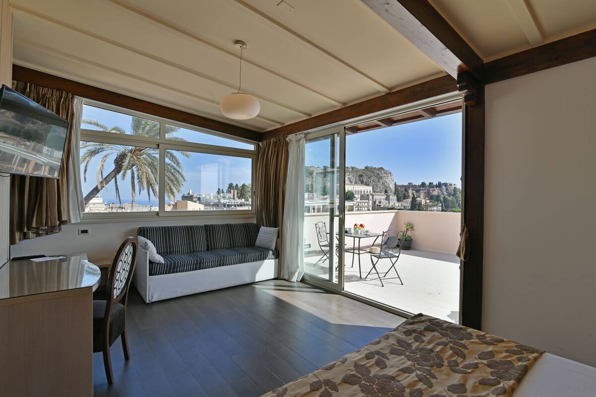 A modern hotel room with a large balcony, a sofa, a dining table, and a view of the city.