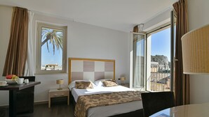 Habitación de hotel con cama, mesita de noche, lámpara y vista a la ciudad a través de un ventanal grande.