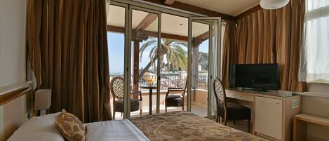 Habitación de hotel con cama, escritorio, televisor y balcón con vista a palmeras y un puente.