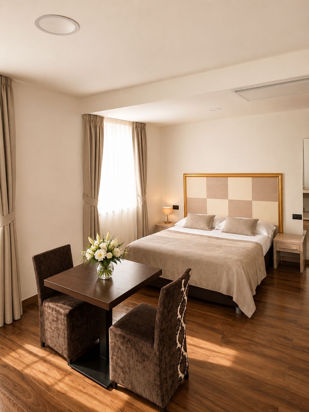 A hotel room with a bed, bedside tables, a desk with chairs, a vase of flowers, and curtains.
